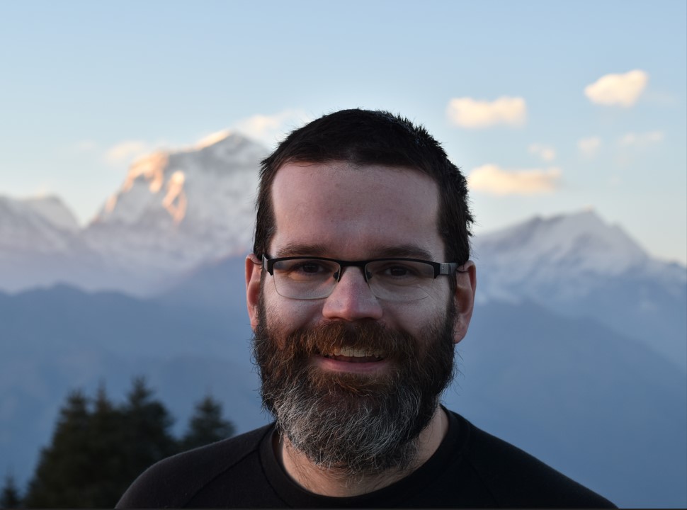 Portrait photograph of Colin Gleason, smiling, in the background there are snow-capped mountains